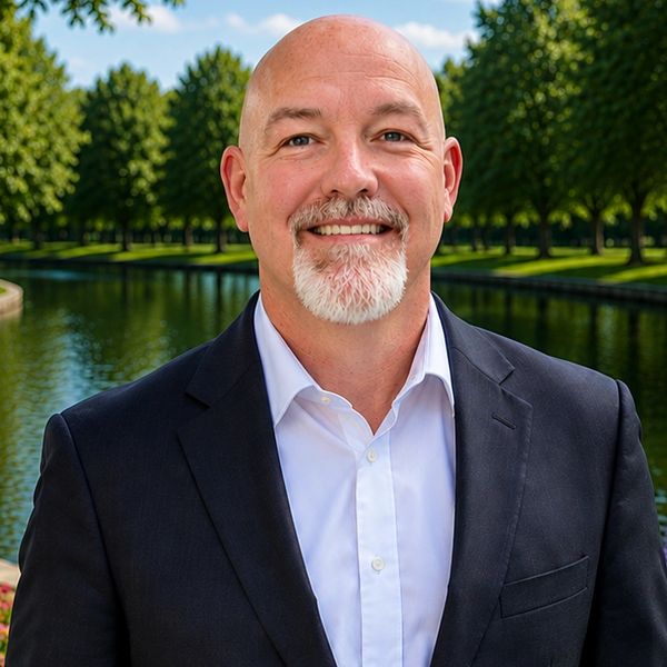 Smiling bald man with a white goatee in a suit by a waterway and trees.