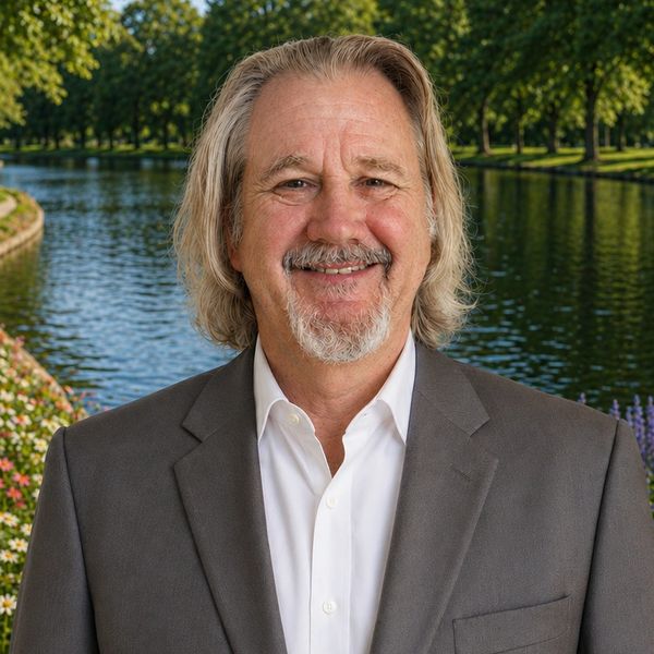 Smiling man in a gray suit stands by a scenic riverside park.