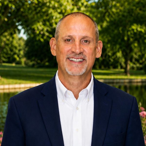 Smiling man in a navy blazer and white shirt outdoors near a pond.