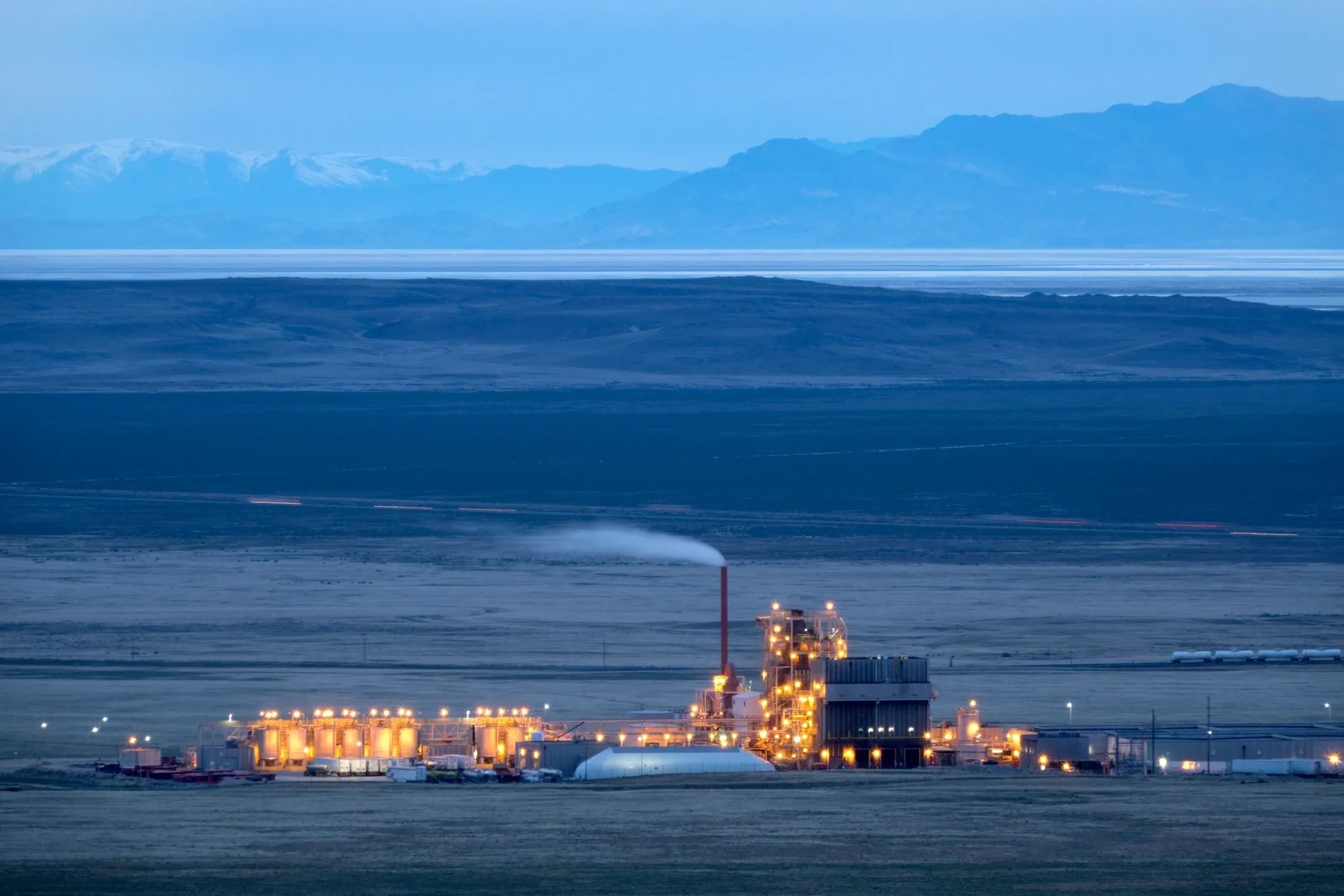Illuminated industrial facility against a mountain backdrop at dusk.
