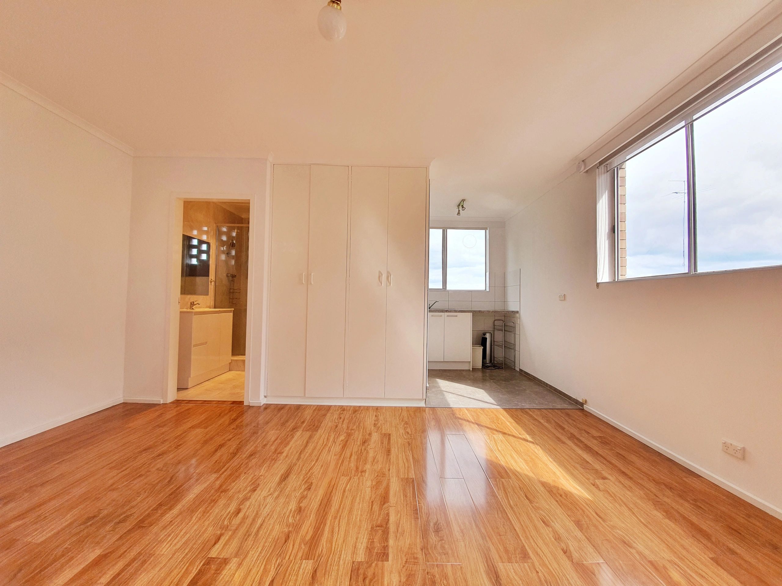 Empty room with wooden floors, white walls, and natural light from a large window.