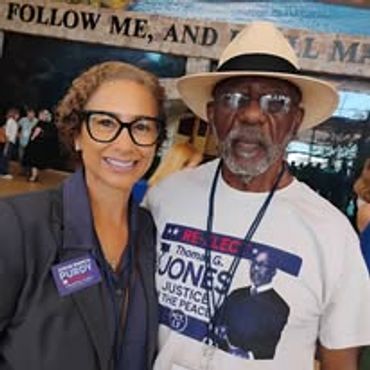Two people smiling, one wearing a justice-themed shirt and the other a blazer with a badge.