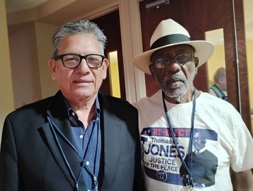 Two elderly men posing indoors, one wearing a suit and glasses, the other in a hat and campaign t-shirt.