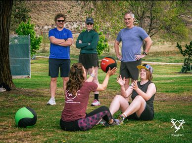 Group exercising outdoors with medicine balls on grass under trees.