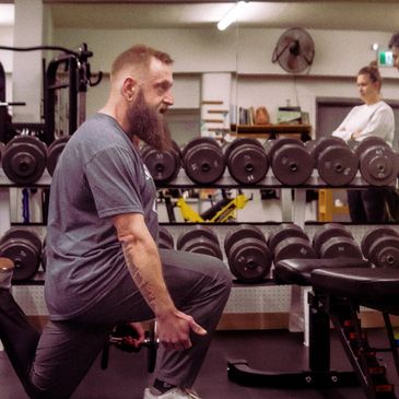 Man with a beard exercising with dumbbells in a gym.