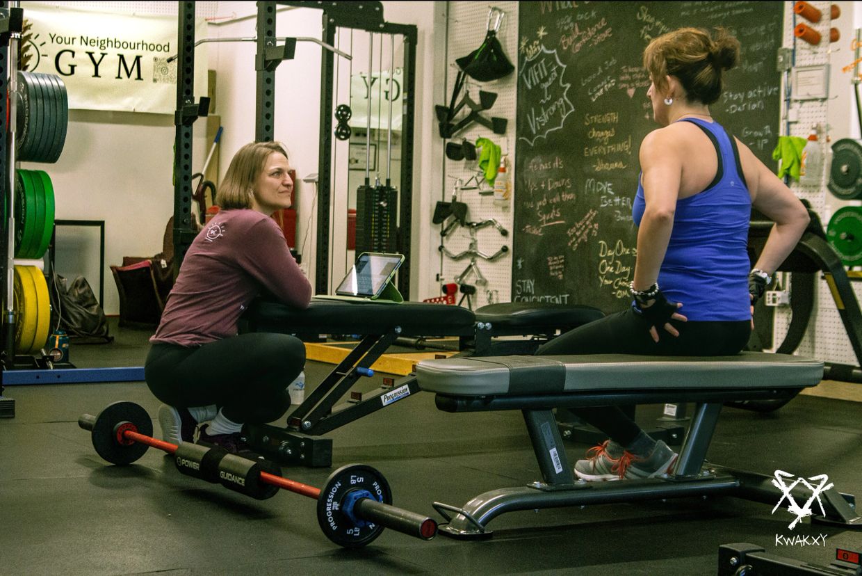 Two women talking in a gym near workout benches and weights.