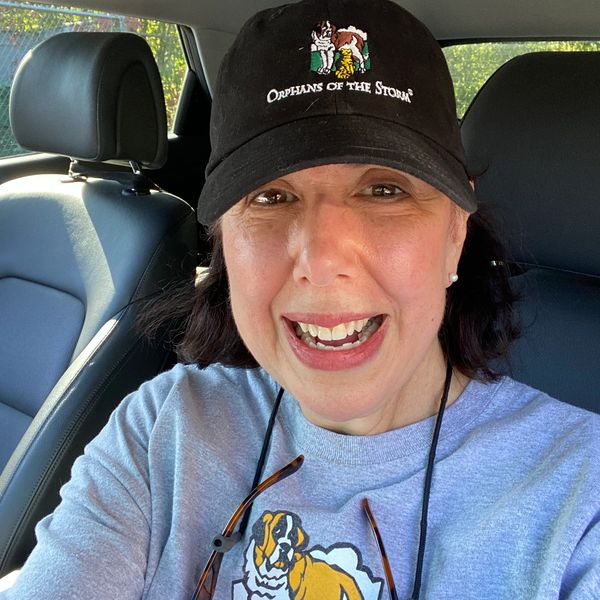 Smiling woman wearing a black cap and gray sweatshirt inside a car.