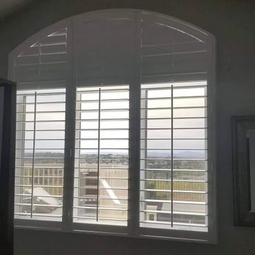 View through white plantation shutters showing outdoor lounge chairs and distant hills.