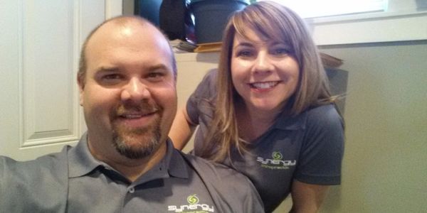 Two people smiling wearing matching Synergy polo shirts indoors.