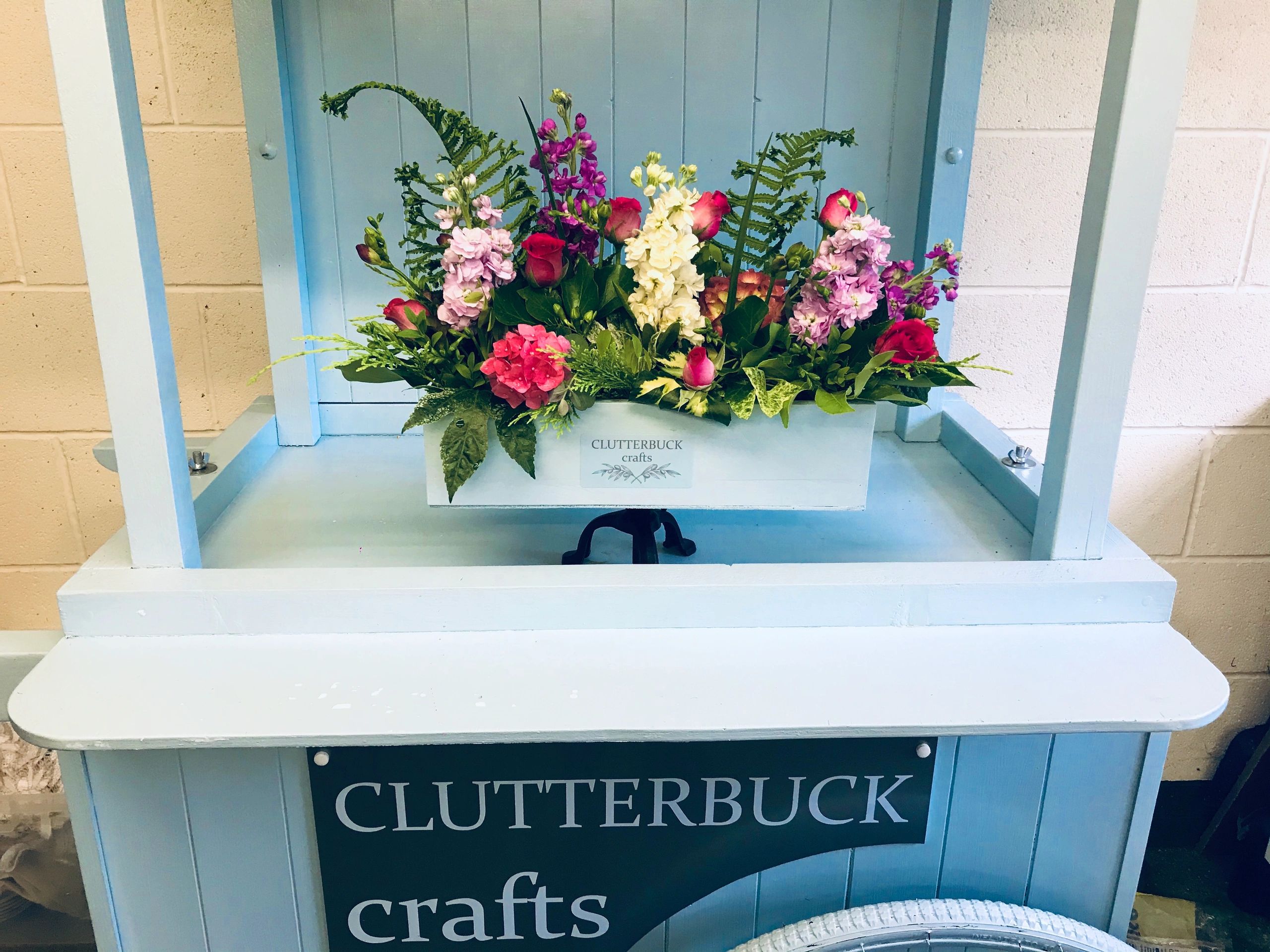 Colorful floral arrangement in a blue wooden cart labeled Clutterbuck crafts.