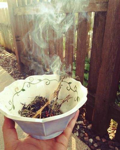 Blue pottery bowl being held by a hand, with incense burning inside, wood fence in background