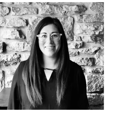 Woman with long hair and glasses smiling against a stone wall.