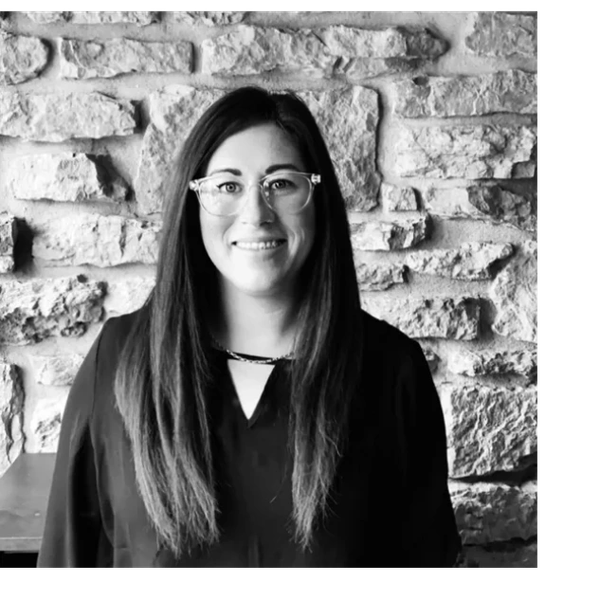 Woman with long hair and glasses smiling against a stone wall.