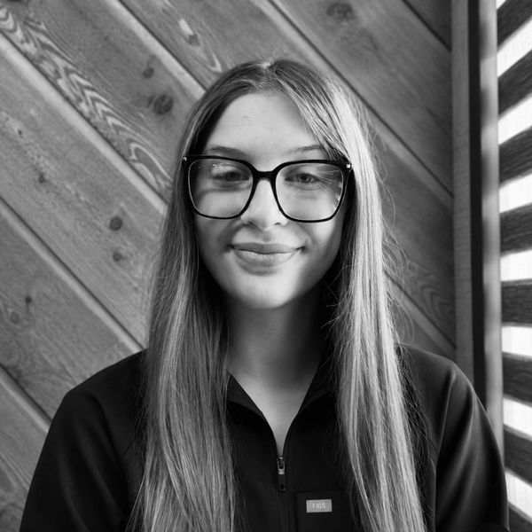 Young woman with long hair and glasses smiling against a wooden background.