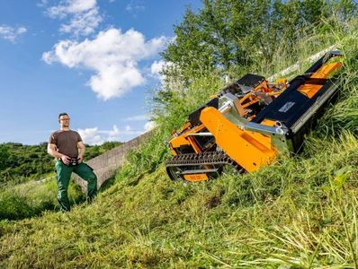 Man operates an orange remote-controlled slope mower on a grassy hillside.