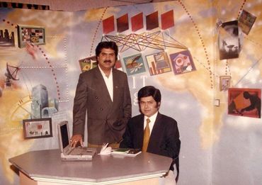 Two men in suits pose in a retro office with a laptop and colorful wall graphics.
