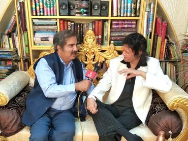 Two men sitting on a golden sofa engaged in an interview amid bookshelves.
