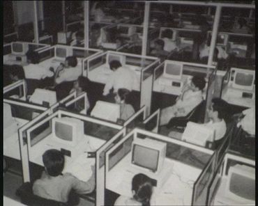Black and white photo of a vintage computer lab with people working in cubicles.