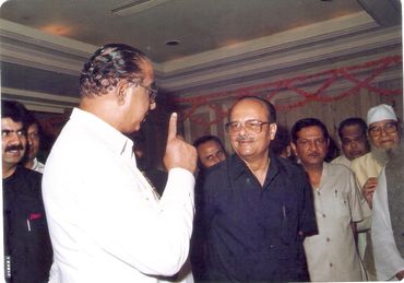 Two men engaged in conversation at a formal indoor event with others around them.