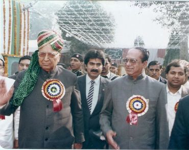 Two elderly men wearing badges at a formal outdoor event with several others around.