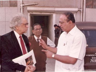 Three men engaged in a serious conversation outside a building, one holding documents.