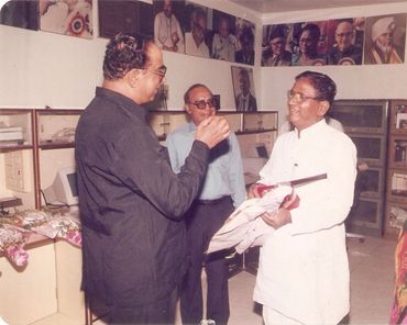 Three men engaged in a conversation in an office decorated with portraits and flowers.