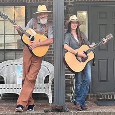 Two people playing guitars on a porch, dressed in casual country-style clothing.