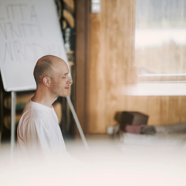 A man in a white shirt meditates indoors near a window and whiteboard.