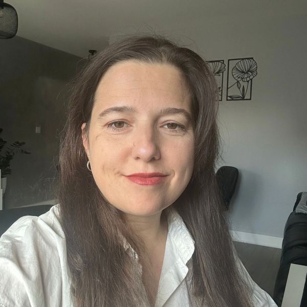 A woman with long brown hair smiles gently, wearing a white shirt indoors.