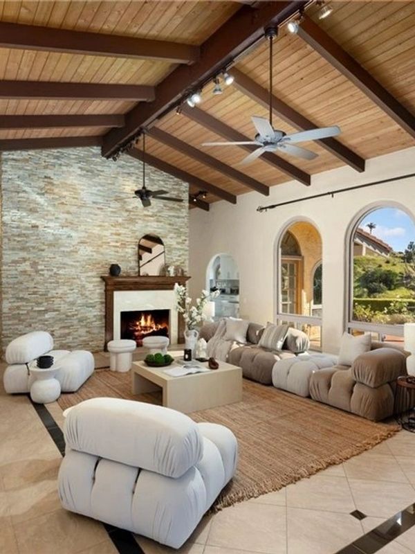 Spacious living room with modern white cushioned furniture and a stone fireplace under a wooden ceiling.