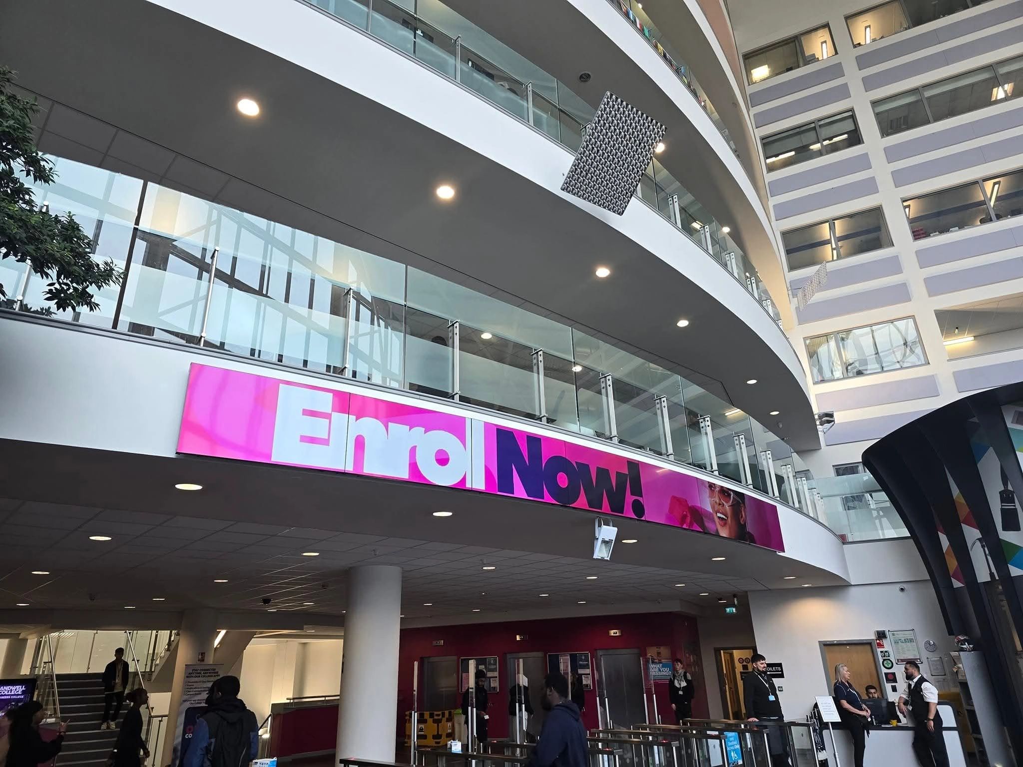 Modern college lobby with a bright 'Enroll Now!' sign and people walking inside.