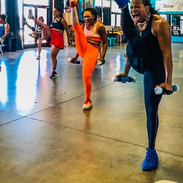 Women energetically exercising with high kicks and dumbbells in a gym.