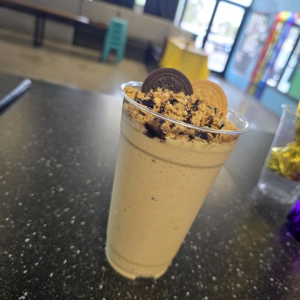 A creamy milkshake topped with crushed cookies and chocolate syrup in a clear plastic cup.