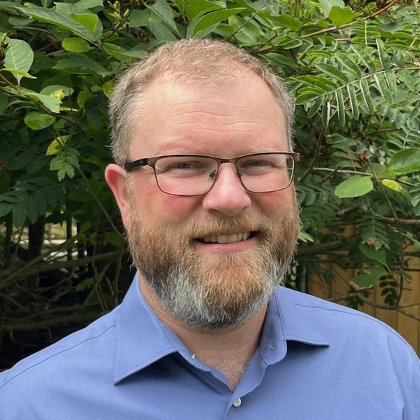 Smiling man with glasses and beard in a blue shirt outdoors.