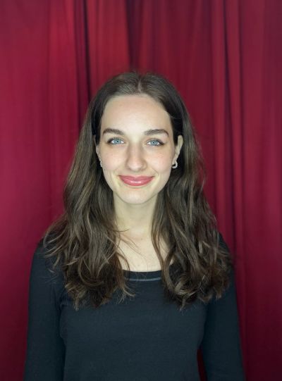 Young woman with long brown hair and blue eyes smiling against a red curtain.