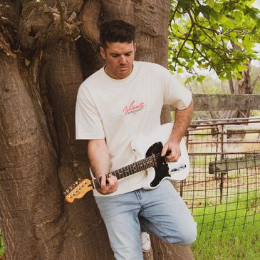 Man playing electric guitar outdoors leaning on a tree.