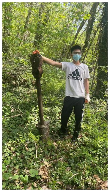 Young man in mask stands in forest holding large rusty old gear mechanism.