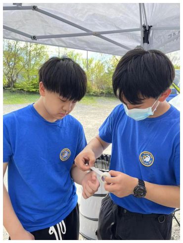 Two boys in blue shirts outdoors, one applying a bandage to the other's arm.