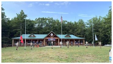 A group of people gathered outside a camp lodge with flags and forest background.