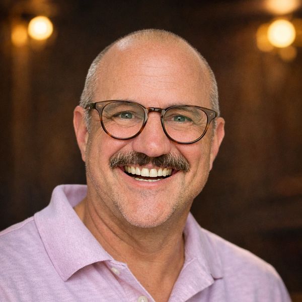 Smiling bald man with glasses and a mustache in a pink shirt.