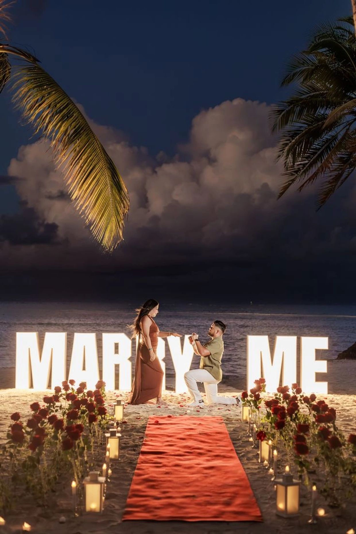 Man proposes to woman on a romantic beach with "Marry Me" lights.