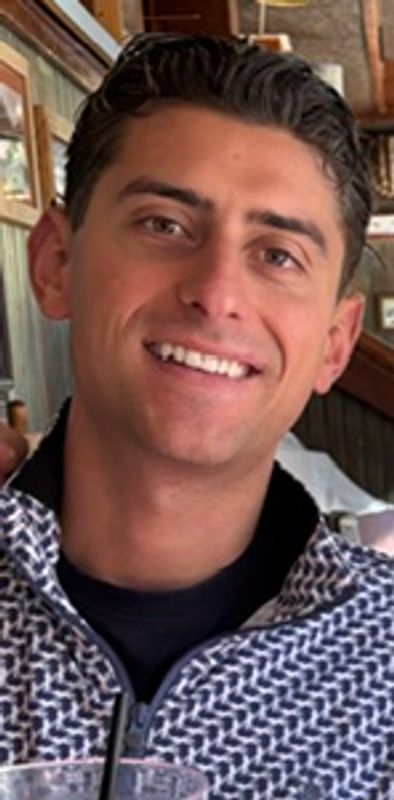 A smiling man with short dark hair in a patterned jacket indoors.
