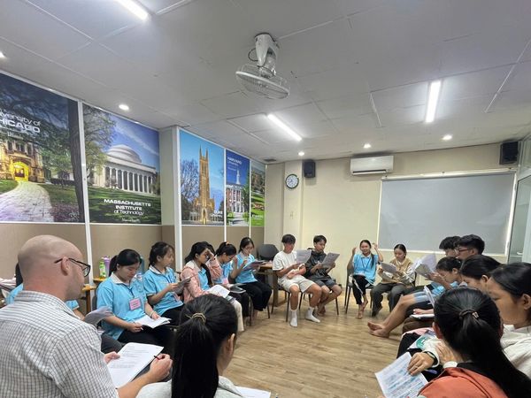 Group of students and teacher in a circle reading documents in a classroom.