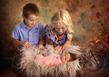 Young brother and sister gaze at newborn baby sister lying on gray fur in antique carriage