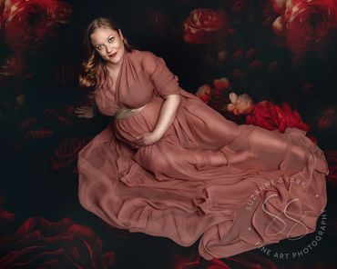 An expectant mother sitting on a backdrop roses with rose colored gown draping around her