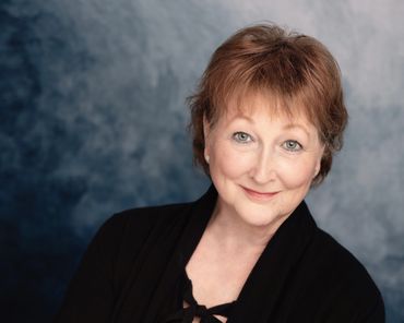 Headshot of older woman with short red hair and black jacket in front of blue backdrop