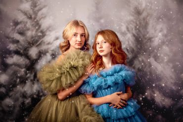 Painterly close-up portrait of blonde and red-haired sisters in fluffy couture gowns in snowy woods