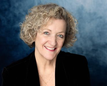 Headshot of blonde older woman in black jacket in front of blue backdrip