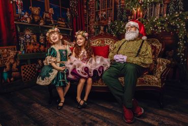 Santa in his Christmas toy shop sits on Victorian sofa with 2 laughing sisters dressed in green, gol
