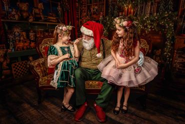 Santa sits in his Toy Shop with 2 little sisters dressed up in Christmas dresses on either side of h